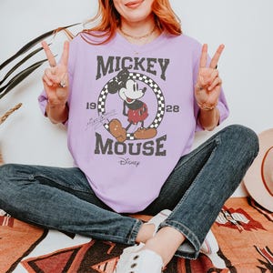 May include: A light purple t-shirt featuring a vintage Mickey Mouse graphic. The design includes Mickey Mouse in a red shorts, a checkered circle, and the text "Mickey Mouse" and "1928".