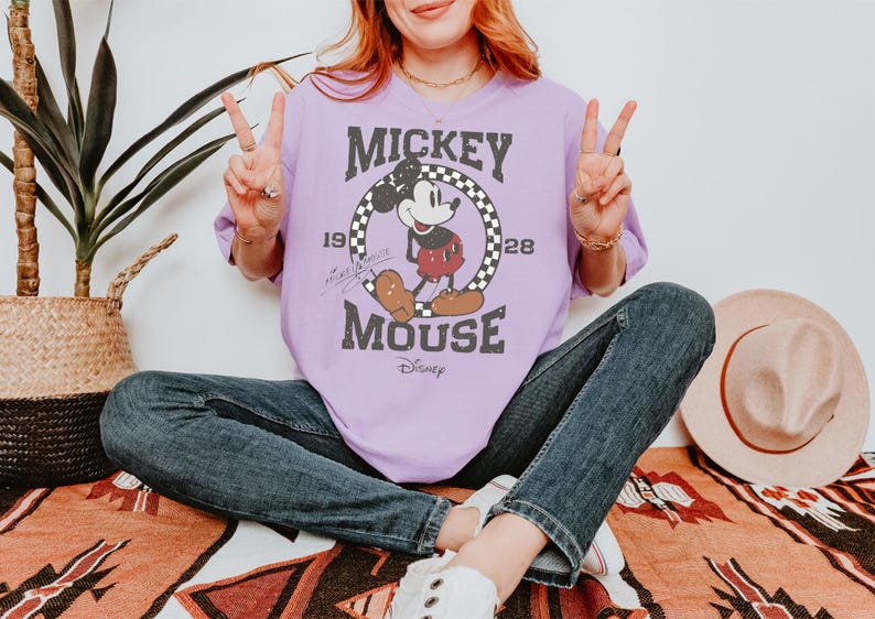May include: A light purple t-shirt featuring a vintage Mickey Mouse graphic. The design includes Mickey Mouse in a red shorts, a checkered circle, and the text "Mickey Mouse" and "1928".