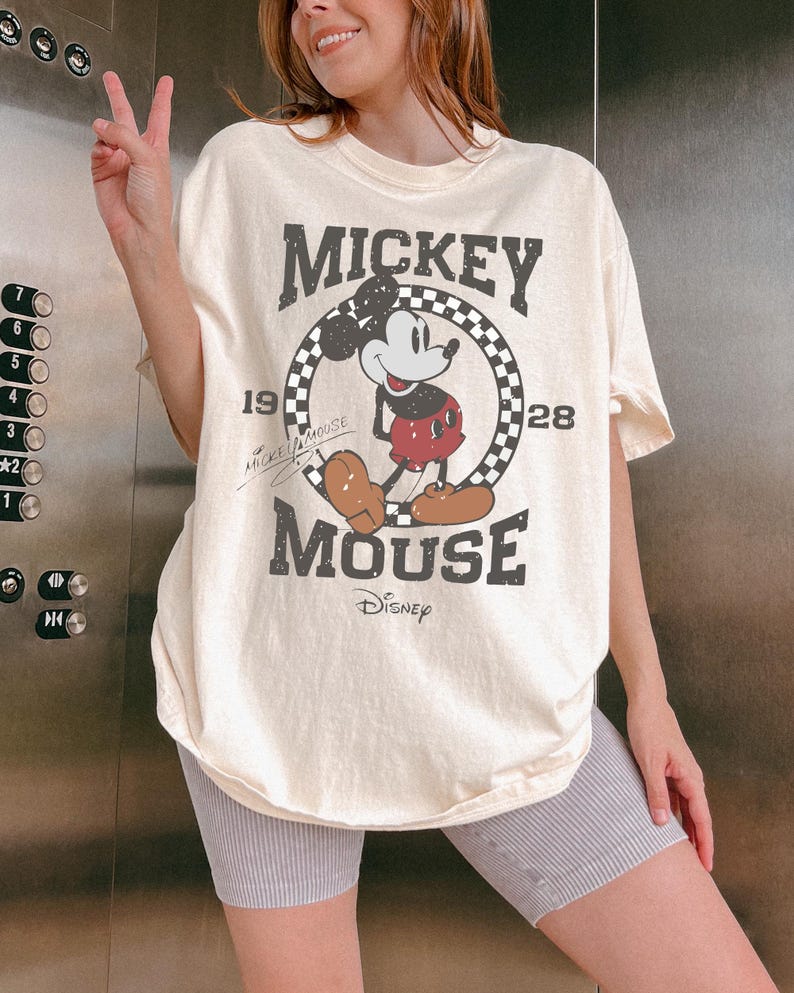 May include: Cream-colored t-shirt featuring a distressed graphic of Mickey Mouse. The design includes the text "MICKEY MOUSE" and "1928" within a checkered circle, with the Disney logo below. The shirt is a relaxed fit.