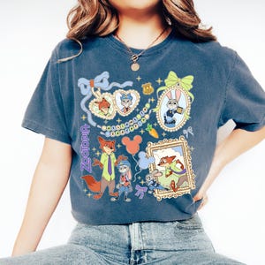 May include: A navy blue t-shirt featuring cartoon characters from the movie Zootopia. The shirt has multiple framed images of the characters, including Nick Wilde and Judy Hopps, with the word "Zootopia" printed vertically on the left side.