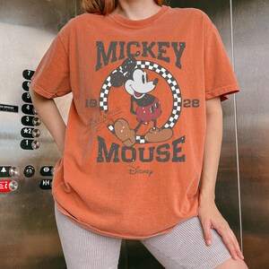 May include: Orange t-shirt featuring Mickey Mouse. The graphic includes Mickey Mouse in a circle with a checkered border, the words "MICKEY MOUSE" and "Disney". The shirt has short sleeves and a vintage look.