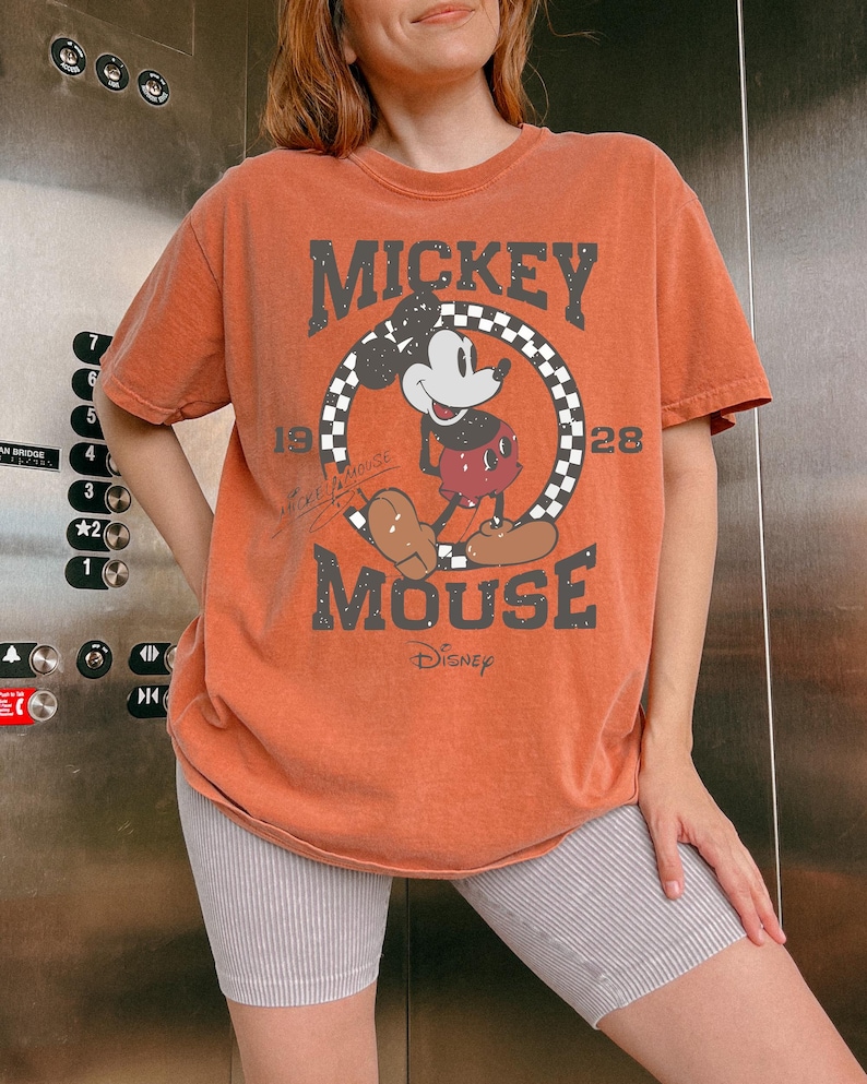 May include: Orange t-shirt featuring Mickey Mouse. The graphic includes Mickey Mouse in a circle with a checkered border, the words "MICKEY MOUSE" and "Disney". The shirt has short sleeves and a vintage look.