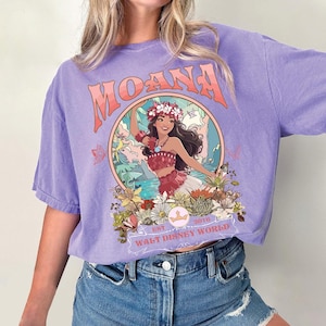 May include: A lilac-colored t-shirt featuring a graphic of a woman in a floral crown, surrounded by flowers and foliage. The word "MOANA" is arched across the top, with "EST 2016 WALT DISNEY WORLD" below the image.