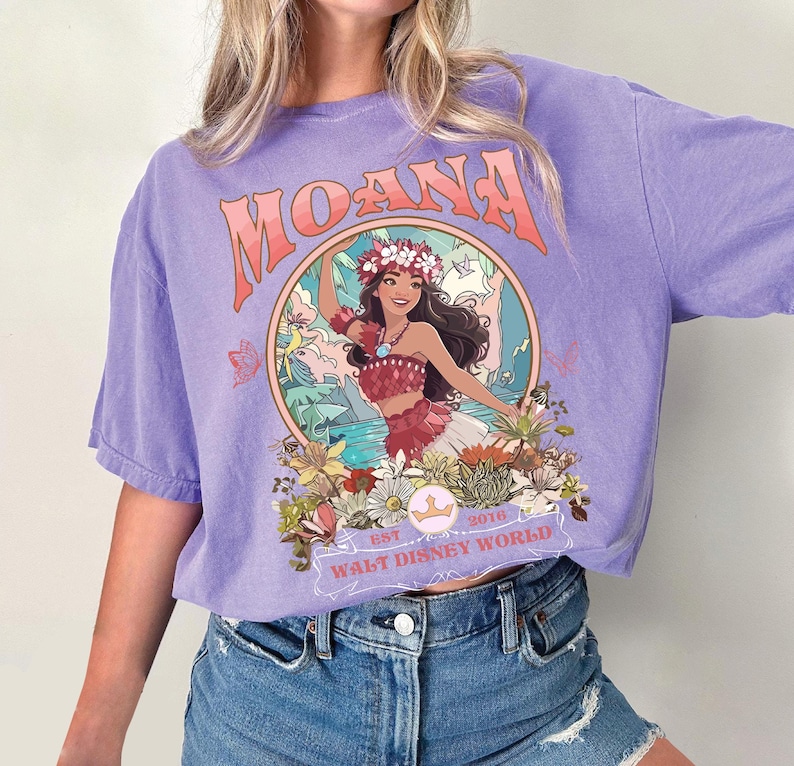 May include: A lilac-colored t-shirt featuring a graphic of a woman in a floral crown, surrounded by flowers and foliage. The word "MOANA" is arched across the top, with "EST 2016 WALT DISNEY WORLD" below the image.