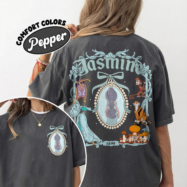 May include: Dark gray t-shirt with a graphic of Jasmine from Aladdin. The design includes the name "Jasmine" in a teal script, a portrait of Jasmine, and other characters. The shirt also has the text "Comfort Colors Pepper".