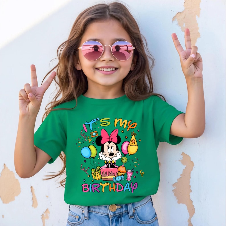 May include: A green t-shirt with the text "It's my birthday" and a graphic of Minnie Mouse. The graphic includes balloons, a cake, and party decorations. The person in the image is wearing pink sunglasses and making a peace sign.