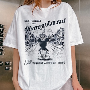 May include: White t-shirt featuring a black and white graphic of Mickey Mouse in front of a Disneyland scene. The text "CALIFORNIA Est. 1955 Disneyland" is above the image, with "The happiest place on earth" below.