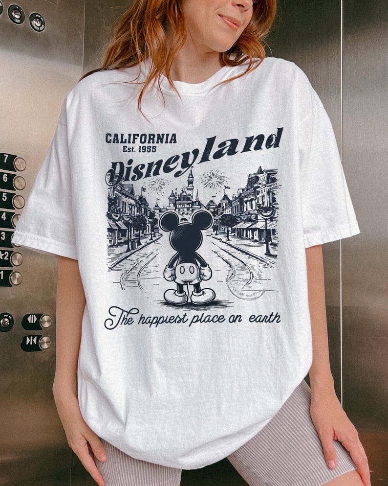 May include: White t-shirt featuring a black and white graphic of Mickey Mouse in front of a Disneyland scene. The text "CALIFORNIA Est. 1955 Disneyland" is above the image, with "The happiest place on earth" below.
