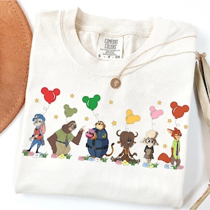 以下が含まれることがあります： クリーム色のComfort Colors Tシャツ。ズートピアのキャラクターがミッキーマウス型の風船を持っている漫画のデザイン。デザインには、ジュディ・ホップス、フラッシュ、クラウハウザー、ニック・ワイルドが含まれています。金のネックレスがシャツの上にかけられています。