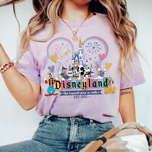 Może przedstawiać: Jasnofioletowa koszulka z logo Disneyland, postaciami Disneya, zamkiem, fajerwerkami i napisem "The happiest place on earth - EST. 1955."