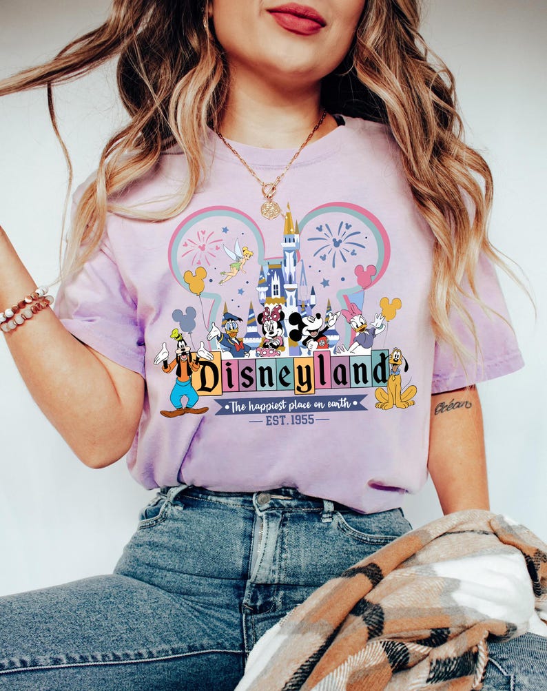 Może przedstawiać: Jasnofioletowa koszulka z logo Disneyland, postaciami Disneya, zamkiem, fajerwerkami i napisem "The happiest place on earth - EST. 1955."