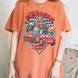 Puede incluir: Camiseta naranja con un dise&ntilde;o de Rayo McQueen y otros personajes de Cars. El dise&ntilde;o incluye el texto "lightning McQueen" y "Cars" sobre un fondo de bandera a cuadros. La camiseta es de corte holgado.