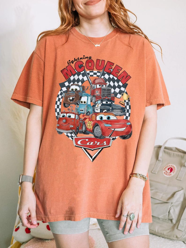 Puede incluir: Camiseta naranja con un dise&ntilde;o de Rayo McQueen y otros personajes de Cars. El dise&ntilde;o incluye el texto "lightning McQueen" y "Cars" sobre un fondo de bandera a cuadros. La camiseta es de corte holgado.