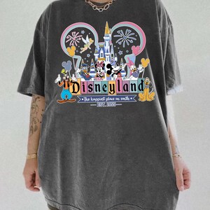 Może przedstawiać: Ciemnoszara koszulka z kolorową grafiką Disneylandu. Projekt zawiera zamek, fajerwerki i r&oacute;żne postacie Disneya. Tekst "Disneyland" jest wyraźnie widoczny, wraz z "The happiest place on earth - EST. 1955."
