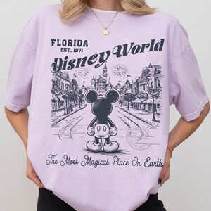 Puede incluir: Una camiseta de color lila claro con un gr&aacute;fico de Mickey Mouse frente a una escena de Disney World. El texto de la camiseta dice "FLORIDA EST. 1971 Disney World" y "The Most Magical Place On Earth."