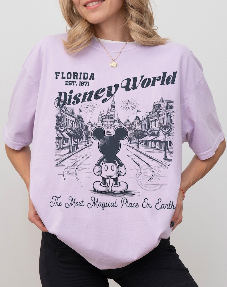 Puede incluir: Una camiseta de color lila claro con un gr&aacute;fico de Mickey Mouse frente a una escena de Disney World. El texto de la camiseta dice "FLORIDA EST. 1971 Disney World" y "The Most Magical Place On Earth."