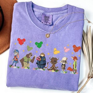 以下が含まれることがあります： 風船を持ったキャラクターの漫画デザインが特徴の紫色のコンフォートカラーズTシャツ。デザインには、ウサギ、ナマケモノ、トラ、バッファロー、羊、キツネが含まれています。金のネックレスと茶色の帽子も表示されています。