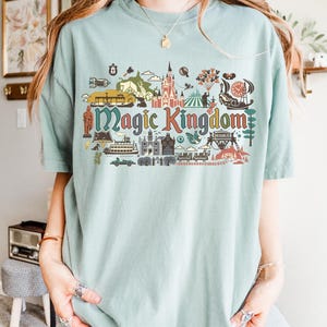 May include: A light blue t-shirt featuring a colorful graphic of the Magic Kingdom. The graphic includes illustrations of a castle, a Ferris wheel, and other theme park elements. The words "Magic Kingdom" are prominently displayed in a multi-colored font.