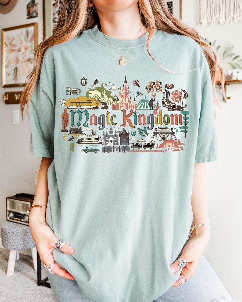 May include: A light blue t-shirt featuring a colorful graphic of the Magic Kingdom. The graphic includes illustrations of a castle, a Ferris wheel, and other theme park elements. The words "Magic Kingdom" are prominently displayed in a multi-colored font.