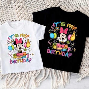 May include: Two children's t-shirts, one white and one black, featuring a colorful graphic of Minnie Mouse. The design includes the text "It's my Birthday" with balloons, a cake, and gifts. The shirts are short-sleeved.
