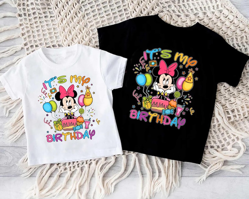 May include: Two children's t-shirts, one white and one black, featuring a colorful graphic of Minnie Mouse. The design includes the text "It's my Birthday" with balloons, a cake, and gifts. The shirts are short-sleeved.