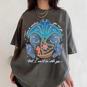 May include: Dark gray t-shirt featuring a colorful graphic design. The design includes a boat, a figure, and stylized waves, with the text "There is nowhere that I won't be with you..."