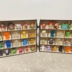 May include: Two wooden display cases with multiple square compartments, each containing a small, colourful plastic character figurine. The collection includes various figures in shades of blue, green, red, yellow, and white, arranged in rows.
