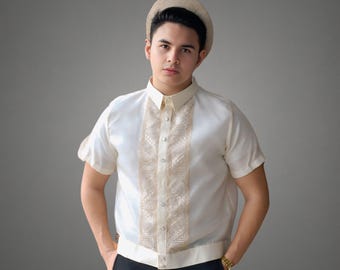 May include: A man wearing a white short-sleeved shirt with a gold embroidered design and a straw hat. He is standing in front of a gray background.