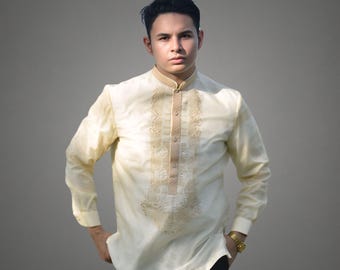 May include: A man wearing a cream-colored long-sleeved shirt with gold embroidery. The shirt has a mandarin collar and a button-up front.