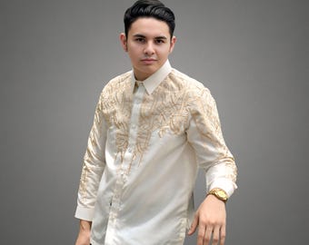May include: A man wearing a white long-sleeved shirt with gold embroidery. The shirt has a button-down collar and a button-up front. The man is wearing a gold watch on his left wrist.