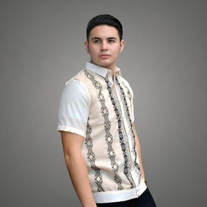 May include: A man wearing a beige short-sleeved shirt with a black and white geometric pattern. The shirt has a button-up collar and a button placket.