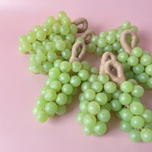 Może przedstawiać: Cztery kiście zielonych winogron z uchwytami z liny na różowym tle. Winogrona są jasnozielone i wydają się gładkie. Uchwyty z liny są jasno brązowe. Obraz jest dobrze oświetlony.