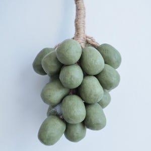 Może przedstawiać: Dekoracyjny pęk zielonych, owalnych przedmiotów, przypominających winogrona, wisi na skręconej linie. Przedmioty mają stonowany zielony kolor i chropowatą powierzchnię. Lina ma naturalny, jasnobrązowy kolor.