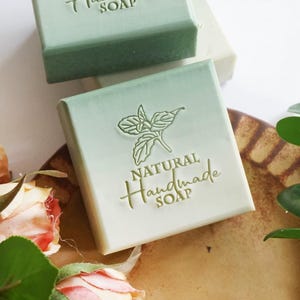 May include: Square, light green bar of handmade soap with the words "NATURAL Handmade SOAP" embossed on the front. A leaf design is above the word "Handmade". The soap is on a brown plate with rose buds.
