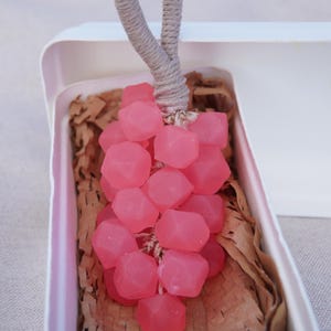 Pode incluir: Um cacho de uvas rosa em forma geométrica com um caule de corda, apresentado numa caixa branca com papel castanho. As uvas são de cor rosa vibrante e o caule é cinzento neutro.