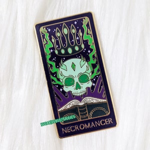 Op de afbeelding: Een gouden en groene emaille pin met een schedel met een kroon en een boek, met de tekst "NECROMANCER" onderaan.