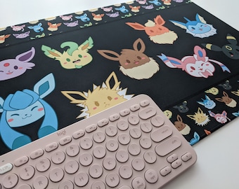 May include: A black mousepad with a repeating pattern of various Pokemon characters. The characters are in a cartoon style and include Eevee, Vaporeon, Jolteon, Flareon, Espeon, Umbreon, Leafeon, Glaceon, and Sylveon. A pink keyboard with round keys is partially visible in the bottom right corner of the image.