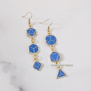 Può includere: Un paio di orecchini pendenti dorati con forme geometriche blu. Gli orecchini sono in metallo e hanno una finitura lucida. Le forme geometriche sono a forma di cubi, diamanti e una piramide. Gli orecchini sono un pezzo di dichiarazione e sarebbero perfetti per un look moderno.