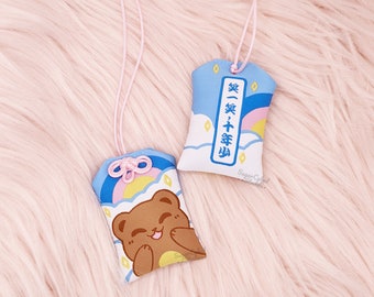 Könnte beinhalten: Zwei kleine, rechteckige Plüsch-Glücksbringer mit rosa Schnur. Einer zeigt einen lächelnden braunen Bären, der andere japanischen Text. Beide haben ein blaues Himmel- und Regenbogen-Design. Die Anhänger liegen auf einer rosa, flauschigen Oberfläche.