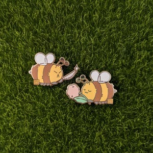May include: Two gold-colored enamel pins of cartoon bees. The bee on the left is holding a sword and has an angry expression. The bee on the right is holding a flower and has a happy expression. Both bees have brown and yellow stripes.