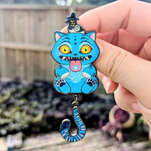Puede incluir: Un pin de esmalte azul con un gato de dibujos animados con ojos amarillos, un sombrero y una cola larga y curvada. El gato tiene la lengua fuera. El pin está sujeto por una mano.