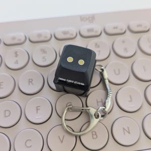 May include: Black keycap keychain with two yellow dots, resembling eyes, and the text "SUGAR CUBED STUDIO" on the side. The keychain is attached to a silver metal ring and chain, and is resting on a pink keyboard.