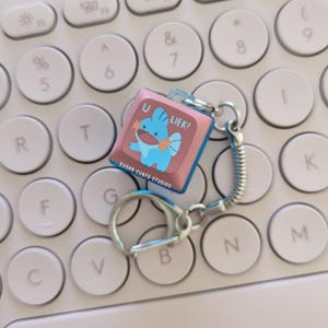 May include: A Mudkip-themed keychain with the text "U LIEK?" on a pink square. The keychain has a silver keyring and a coiled chain, set against a white keyboard. The text "SUGAR CUBED STUDIOS" is also visible.