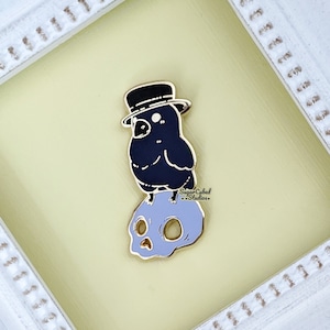 May include: Enamel pin featuring a black crow wearing a top hat, perched atop a light purple skull. The pin has gold-colored trim and the text "Sugar Cubed Studios" is visible. The background is a soft yellow.