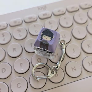 Puede incluir: Un tapón de tecla morado con una ilustración de personaje de dibujos animados de una persona con cabello negro y ojos verdes. El tapón de tecla está unido a un llavero plateado con un pequeño anillo plateado. El tapón de tecla está sobre un teclado rosa.