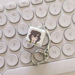 Op de afbeelding: Een witte vierkante sleutelhanger met een cartoonillustratie van een personage met bruin haar en een rood en geel outfit. De sleutelhanger is bevestigd aan een zilveren metalen ketting en sleutelring. De tekst "SUGAR CUBED STUDIOS" is op de sleutelhanger gedrukt. De sleutelhanger ligt op een wit toetsenbord.