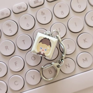 May include: A white keycap keychain with a cartoon illustration of a person with blonde hair wearing a brown hat and a blue jacket. The keychain is attached to a silver chain and a silver ring. The keycap is on a pink keyboard.