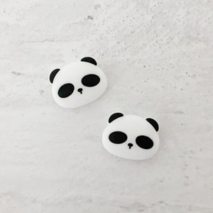 Op de afbeelding: Twee witte pandabeer-gezichtscharms met zwarte ogen en neuzen. De charms zijn gemaakt van glad, wit materiaal en perfect om een ​​toets van whimsy toe te voegen aan elk project.
