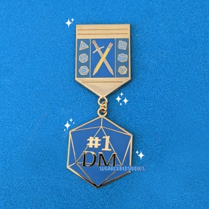 May include: Gold enamel pin featuring a blue and gold die with the text '#1 DM' and the words 'Sugarcubedstudios' at the bottom. The die is attached to a gold enamel pin with a sword and pencil crossed.