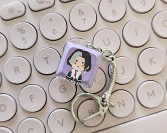 May include: A purple and white keycap keychain with a cartoon character wearing a black and white outfit. The keychain is attached to a silver metal chain and is sitting on a white keyboard.